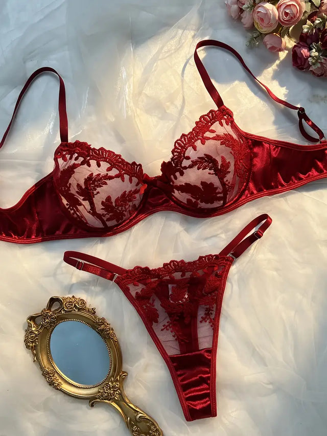 A red lingerie bra set with floral embroidery and lace details, accompanied by a thong with a matching design, displayed on a white surface with a decorative mirror and pink roses in the background.