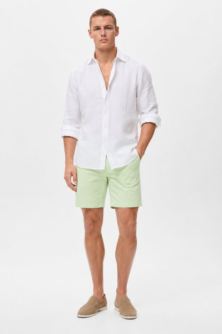 A man modeling a white long-sleeve linen button-down shirt and light green shorts, paired with beige slip-on shoes, against a plain white studio background.